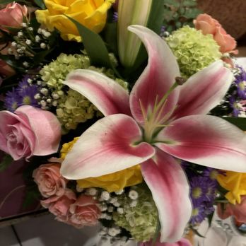 Dawn's Flower Patch - Kiki's flowers - A floral bouquet includes a large pink and white lily at the center, surrounded by yellow roses, pink roses, hydrangeas, and purple flowers. The arrangement features green leaves and filler flowers, set against a blurred background, likely on a table or display area. - Florists Near Me - Wells, Florida