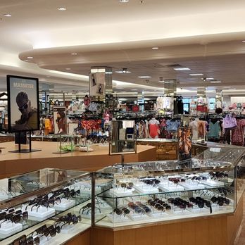 Dillard's - The photo depicts the interior of a department store with a focus on sunglasses displayed in glass cases in the foreground. Behind the cases, clothing racks with various garments, including dresses and tops, are visible. The store has bright lighting and a spacious layout, with additional merchandise and displays further in the background. - Department Stores Near Me - Plantation, Florida