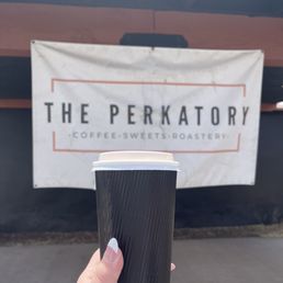 A hand with manicured nails holds a black coffee cup outside The Perkatory coffee shop, with the sign visible in the background.