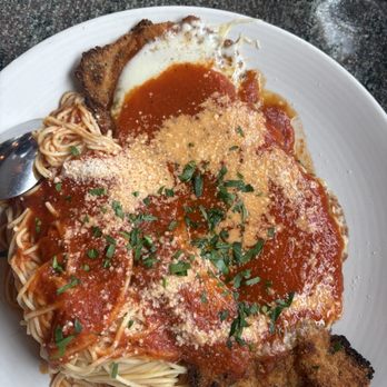 Ciao Amici Restaurant - Chicken parm delicious - A white plate holds a serving of breaded chicken parmesan topped with marinara sauce, grated cheese, and chopped herbs. The dish includes a portion of cooked spaghetti with sauce and a piece of breaded fried item. The plate is set on a dark, speckled countertop, with a spoon and a glass partially visible. - Italian Near Me - Wakefield, Massachusetts