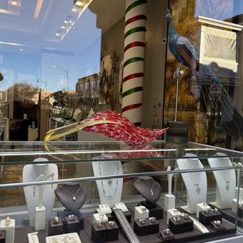 Feathers Galleria - The image shows a jewelry store window display featuring various jewelry items such as necklaces, rings, and pendants arranged on display busts and small stands. A large decorative peacock ornament is positioned behind the jewelry, and a striped pole with red, white, and green stripes is also visible inside the store. The reflection of the street and buildings outside can be seen on the glass window, indicating an outdoor shopping area. - Jewelry Near Me - Santa Fe, New Mexico