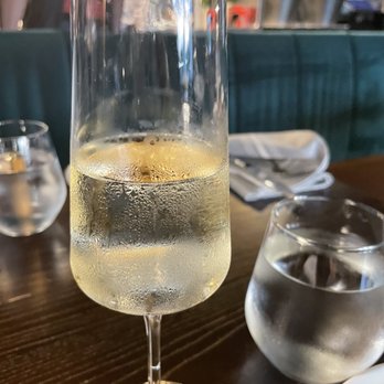 Platea - A tall flute glass filled with sparkling wine or champagne, with condensation on the exterior, on a wooden table in a restaurant setting. - Peruvian Near Me - Pinecrest, Florida
