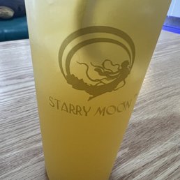 Lemon Kumquat - A yellow plastic cup displaying the Starry Moon logo, which includes a stylized moon with clouds underneath, is positioned on a wooden table. The cup appears to be filled with a beverage and is set in an indoor environment, with part of a plush toy or object visible in the background.