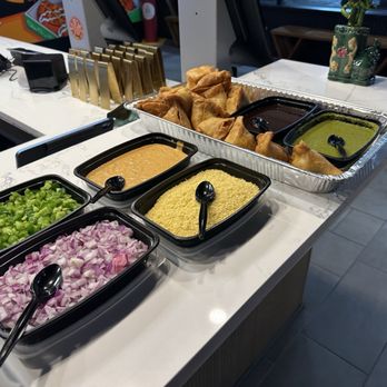 Mazala Pizza - Samosas - A buffet station at Mazala Pizza in Naperville, IL displays samosas in a large aluminum tray, accompanied by three different sauces or chutneys in black containers. In front, there are bowls of chopped green bell peppers, diced onions, and a yellow topping, each with a serving spoon. The background includes a counter with electronic devices and a decorative plant on a white marble surface. The setting appears to be inside a restaurant or food service area. - Chicken Wings Near Me - Naperville, Illinois