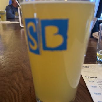 Sandbox Brewhouse - A tall glass filled with a yellow drink featuring a blue and yellow logo, set on a wooden table in a restaurant or bar setting. - Brewpubs Near Me - Frederick, Maryland
