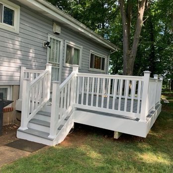 Exterior Experts - Stairs - A white wooden deck with stairs and railing is attached to a gray house with multiple windows, surrounded by green trees and grass. - Siding Near Me - Baltimore, Maryland