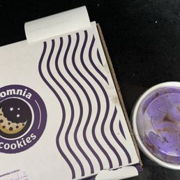 A white box with purple wavy lines and the Insomnia Cookies logo, featuring a moon and stars, is positioned on a black surface. Next to it is a white cup containing purple ice cream, with some melted portions visible on the surface. The image suggests a snack or dessert setting at Insomnia Cookies.