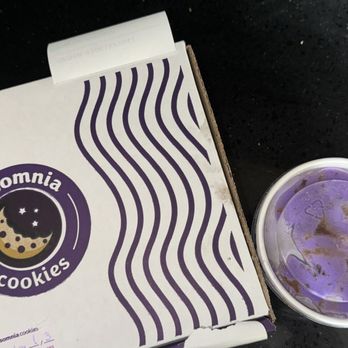 Insomnia Cookies - A white box with purple wavy lines and the Insomnia Cookies logo, featuring a moon and stars, is positioned on a black surface. Next to it is a white cup containing purple ice cream, with some melted portions visible on the surface. The image suggests a snack or dessert setting at Insomnia Cookies. - Bakeries Near Me - Naperville, Illinois