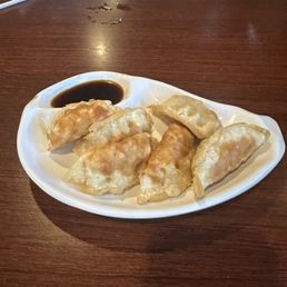 Gyoza - A white, oval-shaped plate holds several fried dumplings with a golden-brown, crispy exterior. A small compartment in the plate contains soy sauce for dipping. The plate is positioned on a dark wooden table, with some water or oil droplets visible nearby. The setting appears to be a restaurant or casual dining area.