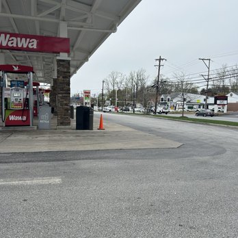 Wawa - Prices, right corner! - The image shows the exterior of a Wawa convenience store with a gas station area. The store has a canopy with the Wawa logo, and there are gas pumps visible on the left side. The parking lot and street are seen in the background, with several cars and utility poles. The scene appears to be on a cloudy day with minimal activity. - Convenience Stores Near Me - Upper Providence, Pennsylvania