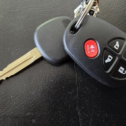 Key & Remote - A metal car key with a plastic head is attached to a black remote fob with four buttons: lock, unlock, trunk release, and panic alarm. The remote is connected to a small metal keyring, and the surface beneath is black and textured.