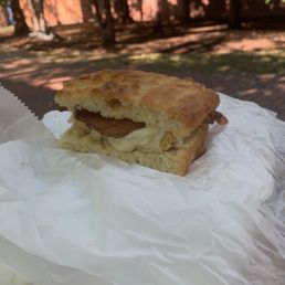 Breakfast Sandwich - A breakfast sandwich featuring bacon, egg, and cheese is shown on crumpled white paper outdoors. The sandwich is made with toasted bread, and the filling includes crispy bacon, a cooked egg, and melted cheese. The background includes trees and a brick wall, indicating an outdoor setting with natural light.