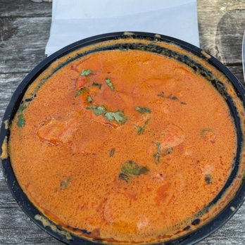 Karam Restaurant - a plate of food on a picnic table - A black plastic bowl holds a serving of Indian-style curry with a reddish-orange color, garnished with cilantro leaves. The bowl is placed on a rustic wooden table, with some food residue visible on the rim. In the background, there is a white paper sheet and a partially visible container with grilled food, indicating an outdoor dining setting. - Indian Near Me - Schenectady, New York