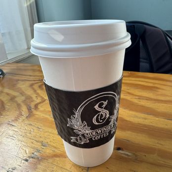 Sidewinder Coffee - Coffee - A white disposable coffee cup with a black Sidewinder Coffee sleeve is placed on a wooden table, with a window and a backpack in the background. - Coffee & Tea Near Me - Cincinnati, Ohio