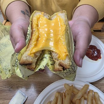 The Hat - Look at that cheese pull! - A person wearing a yellow hoodie is holding a cheeseburger with melted cheese, partially wrapped in yellow paper, above a plate of French fries and a small dish of ketchup. The burger appears to have a toasted bun and a slice of melted cheese. The setting is a wooden table with food items arranged on white disposable plates. - American Near Me - Pasadena, California