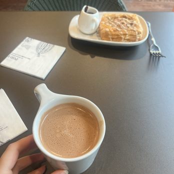 Hotel Chocolat - Drinking chocolate - A person holds a mug of hot chocolate at a table with a pastry and a small coffee pot in the background. - Chocolatiers & Shops Near Me - Chicago, Illinois