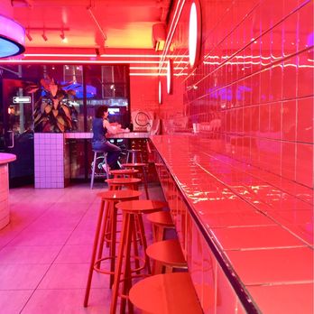 Moonburger - Cool Vibes! - The image shows the interior of a contemporary restaurant or bar with vibrant pink and red lighting. The right side features a long tiled counter with matching red stools arranged along it. The wall behind the counter is covered with glossy red tiles, reflecting the lighting. In the background, a person is seated at the counter, facing away, with a large window or glass door behind them, revealing a plant and city lights outside. The ceiling has exposed pipes and track lighting, contributing to an industrial aesthetic. - Burgers Near Me - Brooklyn, New York