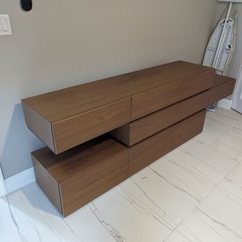 Pure Moving - A wooden sideboard with a sleek, minimalist design and multiple drawers is placed against a light-colored wall in a room. The floor features large, light-colored tiles with subtle veining. An ironing board is stored upright in the corner, and a small device is mounted on the wall nearby. - Movers Near Me - Miami, Florida