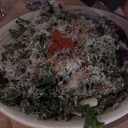 Swiss Chard Caesar Salad - A large bowl contains a Caesar salad with leafy greens, grated cheese, sunflower seeds, and a small amount of red roe placed on top. A glass of water is visible in the background, along with a dark-colored object, possibly a person's arm or clothing. The setting appears to be a dining table in a restaurant or casual dining environment.