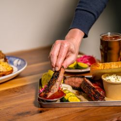 Founding Farmers & Distillers - A hand reaches in to pick up a glazed spare rib from a metal BBQ tray loaded with sliced smoked brisket, bread and butter pic...