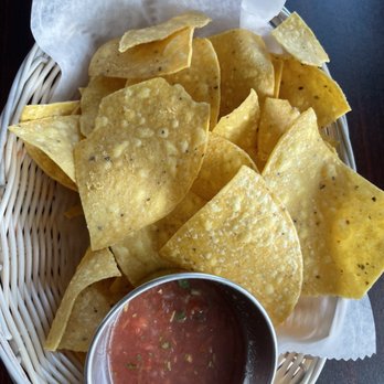 Estefani's Restaurant - Chips & salsa - A woven basket contains numerous golden, crispy tortilla chips with some broken pieces, lined with white parchment paper. A small metal cup of red salsa with visible herbs and chopped vegetables is placed in front of the chips. The basket is on a dark-colored table surface, suggesting a casual dining setting. - Breakfast & Brunch Near Me - Lynn, Massachusetts