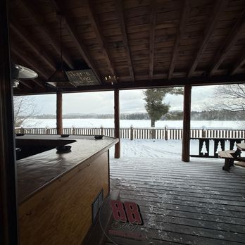 Daikers - A covered outdoor deck with a wooden ceiling and floor, featuring a railing with decorative cutouts. The deck overlooks a snow-covered landscape with a lake and trees in the background under a cloudy sky. The scene suggests a winter setting at a lakeside location. - New American Near Me - Old Forge, New York
