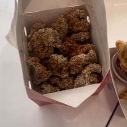 A paper container filled with fried chicken pieces is positioned on a white surface. The chicken appears crispy and seasoned. Part of a drink in a plastic cup and another food item are visible to the right. The image is taken from above, showing the food presentation and setting.