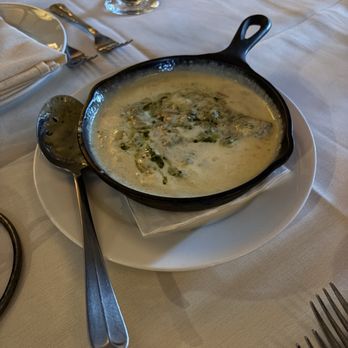 Roaring Fork - Queso Puerco - A bowl of creamy green soup, likely made with herbs or spinach, is presented in a black cast iron skillet placed on a white plate. The table setting includes a spoon, a fork, a napkin, and a wine glass, indicating a dining environment. The tablecloth is white, and the background shows additional tableware and a glass of dark beverage. - American Near Me - Austin, Texas