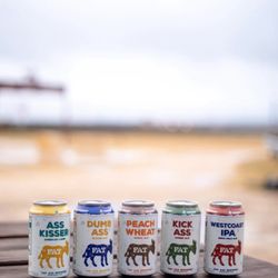 Fat Ass Brewery - Lineup of Fat Ass Ranch beer cans including Ass Kisser, Dumb Ass, Peach Wheat, Kick Ass, and West Coast IPA on an outdoor table