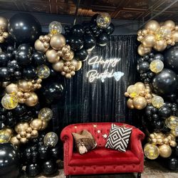 Birthday party setup with black and gold balloon arch, red velvet loveseat, "Happy Birthday" neon sign, and elegant event backdrop