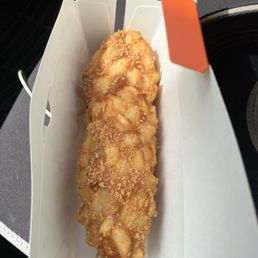 Hot Dog - A deep-fried potato dog on a wooden stick is shown inside a white paper tray. The potato dog has a crispy, golden-brown coating with visible potato chunks. The tray is set on a dark surface, possibly a table or counter, with part of a person's clothing visible in the background. The image captures a casual food item from a Korean-style corn dog vendor.