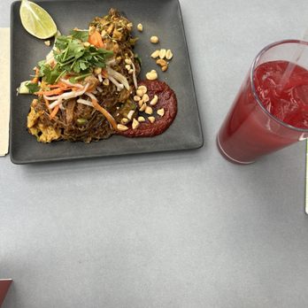 Vital Root - Pad Thai with beet lemonade - A square black plate contains Pad Thai topped with cilantro, shredded vegetables, and peanuts, with a lime wedge on the side. Next to the plate, there is a tall glass filled with beet lemonade, featuring a straw. The meal is set on a gray table, with part of a person's arm visible at the top of the image. - Vegetarian Near Me - Denver, Colorado