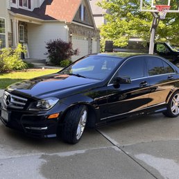 A black Mercedes-Benz sedan with tinted windows is parked in a suburban driveway, with a basketball hoop and a tree in the background.