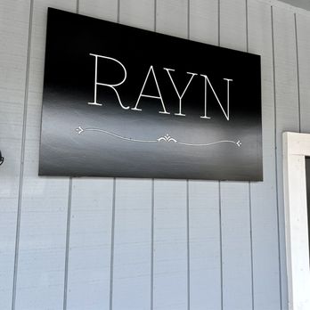 Rayn - A black rectangular sign with white lettering reading "RAYN" is mounted on a gray wooden wall outside a building. - Home Decor Near Me - Temecula, California