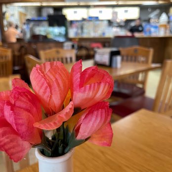 Steamers - Nice tables - A pink artificial flower arrangement in a white vase is positioned on a wooden table inside a restaurant. The background shows wooden chairs, a counter with various items, and blurred figures of people, indicating a casual dining environment. The focus is on the flower arrangement, with the restaurant interior slightly out of focus. - Hot Dogs Near Me - Winfield, Illinois