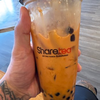 Sharetea - A person holds a large, clear plastic cup of bubble tea with tapioca pearls at Sharetea in Aurora, Illinois, with a wooden table and indoor setting in the background. - Bubble Tea Near Me - Aurora, Illinois