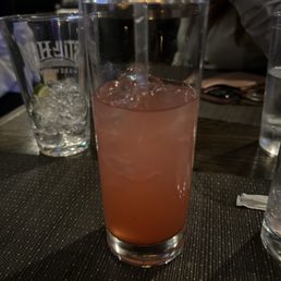 Jared's rum punch - A pink cocktail with ice cubes is served in a tall, clear glass with a straw. The drink appears to be a mixed beverage, possibly a fruit-flavored cocktail, placed on a dark textured table. In the background, there are other glasses, a person wearing a gray shirt, and a dimly lit bar or restaurant setting.