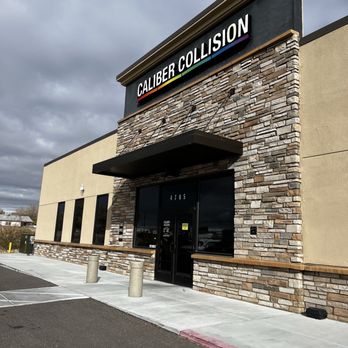 Caliber Collision - The image shows the exterior of Caliber Collision, a business located at 4705. The building has a stone facade around the entrance, large windows on the side, and a black sign with white text displaying the business name. The sidewalk in front is concrete with bollards, and the parking lot is visible with marked spaces. The sky is partly cloudy, and the overall scene is well-lit. - Body Shops Near Me - Rio Rancho, New Mexico