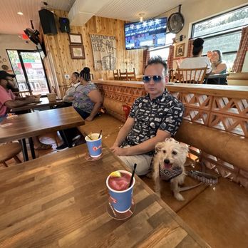 Chalmers - A man wearing sunglasses and a patterned shirt sits at a wooden table with two drinks, accompanied by a small dog. The restaurant has wood-paneled walls, a TV, and several patrons. - Bars Near Me - Austin, Texas