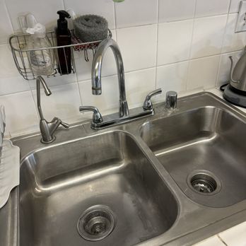 Miraculous Cleaning - Sink and knobs are spotless and shiny - A stainless steel double kitchen sink features a chrome faucet with a soap dispenser and a separate side faucet. Above the sink, a wall-mounted wire rack holds hand soap, a black pump bottle, and a sponge. Decorative hooks with colorful fruit-themed designs are attached to the white tiled wall. To the right, a kettle is partially visible on the countertop. The sink appears clean and well-maintained. - Home Cleaning Near Me - Pasadena, California