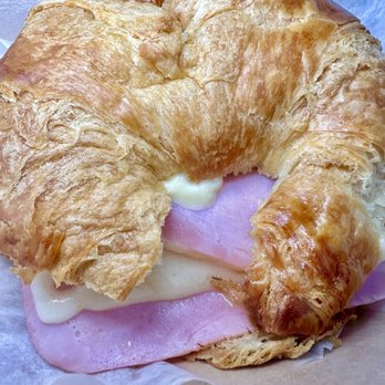 Picnic - Ham and cheese croissant - A close-up image of a croissant sandwich containing slices of ham, melted cheese, and mayonnaise. The croissant has a golden-brown, flaky exterior and is placed on parchment paper. The background features a blue surface, emphasizing the sandwich's details. - Cafes Near Me - Newport, Rhode Island
