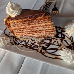 Smith Island Cake - A slice of Smith Island cake, featuring multiple thin layers of cake and filling, topped with a dollop of whipped cream. The cake is served on a rectangular white plate decorated with swirls of chocolate and vanilla sauces, with additional whipped cream dollops around the cake. The presentation is set on a white tablecloth, with utensils visible in the background.