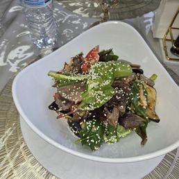 Eggplant Salad - A mixed green salad with tomatoes, eggplant, and sesame seeds is presented in a white bowl on a dining table with water bottles and utensils.