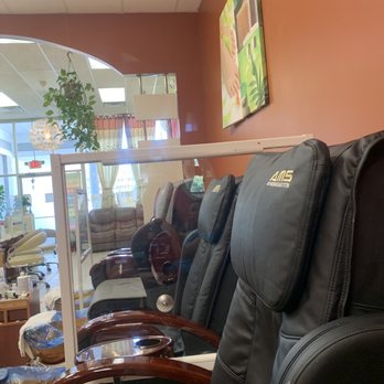 2Q Nails - Pedicure area - The image shows a row of black massage chairs with wooden armrests positioned in a nail salon's pedicure area. Each chair has a headrest with the logo 'AMS' and is separated by clear acrylic dividers. The background includes a wall with a colorful painting, a window with curtains, and some indoor plants, indicating a comfortable and organized salon environment. - Nail Salons Near Me - Ledyard, Connecticut