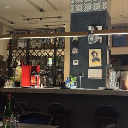 Inside / bar - The image shows the interior of a bar with a counter and seating area. The bar has a decorated wall with artwork, masks, and a thermostat. Behind the counter, shelves hold various bottles of alcohol and bar equipment. The ceiling has track lighting, and there are hanging light fixtures above the bar. The seating area includes chairs and tables with drinks and bottles placed on them. The overall ambiance appears cozy and modern.