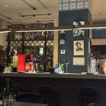 Song E Napule - Inside / bar - The image shows the interior of a bar with a counter and seating area. The bar has a decorated wall with artwork, masks, and a thermostat. Behind the counter, shelves hold various bottles of alcohol and bar equipment. The ceiling has track lighting, and there are hanging light fixtures above the bar. The seating area includes chairs and tables with drinks and bottles placed on them. The overall ambiance appears cozy and modern. - Pizza Near Me - Rutherford, New Jersey