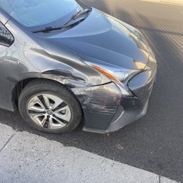 Car - The image depicts a black hybrid car with visible front-end damage, including a dented bumper and fender, parked on the side of a paved road. The car's front left wheel and tire are visible, and the vehicle appears to be stationary. The background includes a shadow cast on the road and part of a sidewalk.