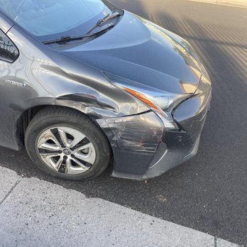 Body Master - Car - The image depicts a black hybrid car with visible front-end damage, including a dented bumper and fender, parked on the side of a paved road. The car's front left wheel and tire are visible, and the vehicle appears to be stationary. The background includes a shadow cast on the road and part of a sidewalk. - Body Shops Near Me - Albuquerque, New Mexico