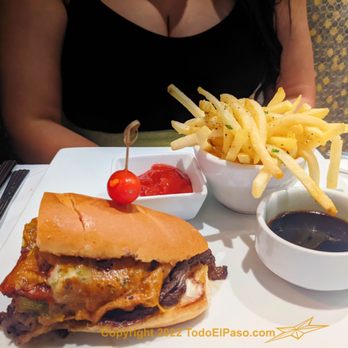 Cafe Central - A woman is seated at a table with a cheeseburger topped with a cherry, French fries in a bowl, ketchup, and a cup of dark soda, with cutlery on the side. - Salad Near Me - El Paso, Texas
