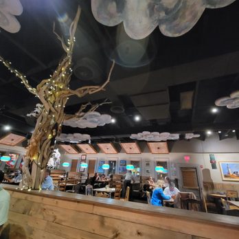 Range Cafe - Inside... Cloudy with a chance of Green Chile.. - The image shows the interior of a restaurant with a dark ceiling decorated with cloud-shaped elements. A wooden partition with a carved, twisted tree sculpture wrapped in string lights is visible in the foreground. The dining area includes wooden tables and chairs, with patrons seated and dining. The walls are decorated with framed pictures and illuminated menu boards, and the overall ambiance is cozy and themed with natural and sky-inspired decor. - Breakfast & Brunch Near Me - Albuquerque, New Mexico